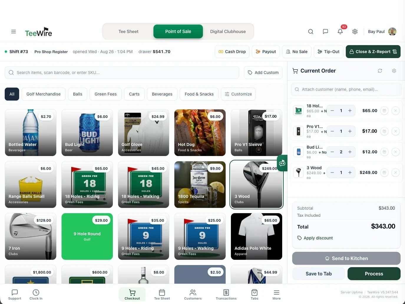 The TeeWire register on the checkout screen, with a grid of priced product tiles covering green fees, balls, clubs, apparel, beverages and food, filtered by category across the top. The current order runs down the right side listing an eighteen hole round, a sleeve of Pro V1s, two Bud Lights and a 3 Wood, with the running subtotal, a tax included line and the total beneath them and the send to kitchen, save to tab and process actions at the foot. The open shift, its drawer total and the cash drop, payout, no sale, tip out and close and Z report controls run across the header.